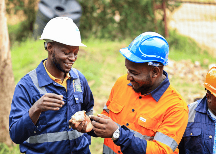 Two smiling workers in hard hats and safety gear examine a rock sample outdoors.